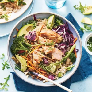 Salmon & Brown Rice Super Bowl – A colorful bowl with grilled salmon, brown rice, broccoli, shredded carrots, red cabbage, and avocado slices on a light blue table.