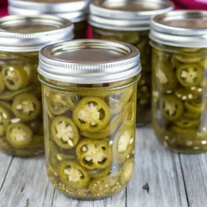 Close-up of mason jars filled with sliced canned jalapeños in brine on a rustic wooden surface.