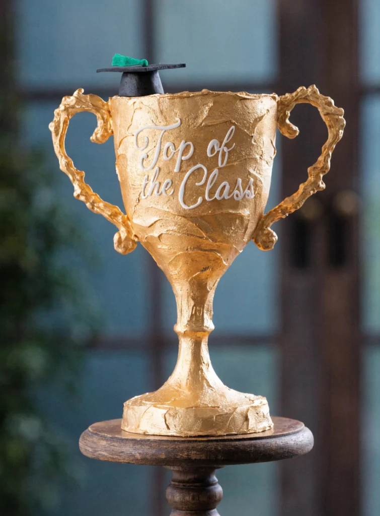 Golden trophy-shaped graduation cake with fondant cap and "Top of the Class" message for kids